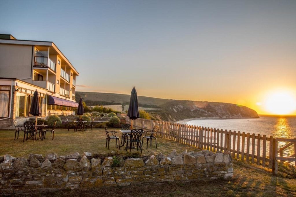 a white coastal hotel building on a low cliff overlooking the sea in swanage dorset ith the sun setting in the background 