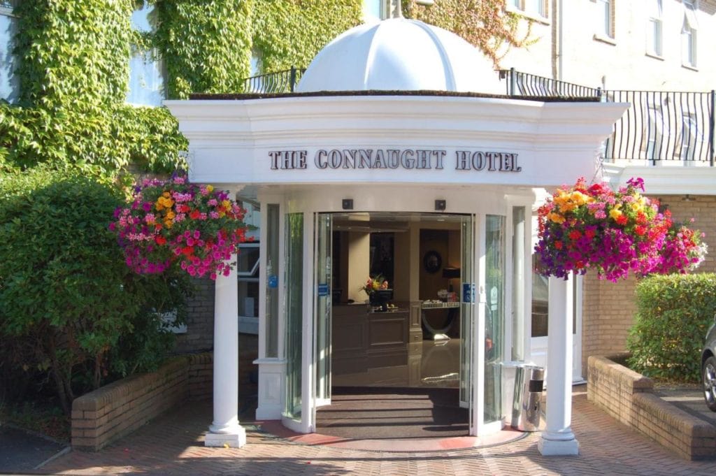 a grand porch entrance with a white domed roof and white pillars. the entrance is attached to a white hotel building covered in green ivy. there are two hanging baskets on either side of the door wih pink flowers. the sign reads The Connaught Hotel. 