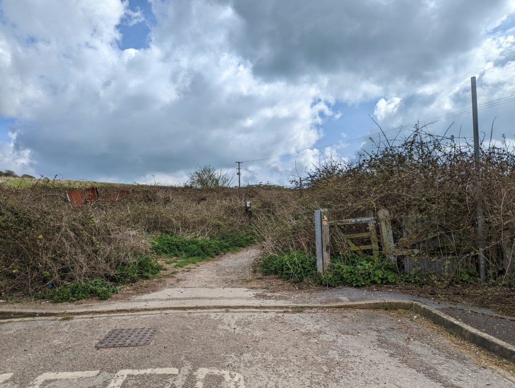 a gravel path leading away from the end of a road between hedgerows with a broken wooden gate to the right