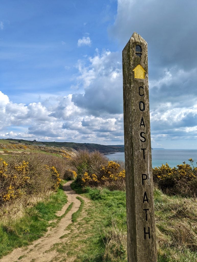 a wooden signpost with a yellow arrow pointing forwards and the words coast path printed on it. the sign is next to a narrow dirt path which leads towards a gap in some yellow gorse bushes. Beyond the bushes are low grassy cliffs and the sea.