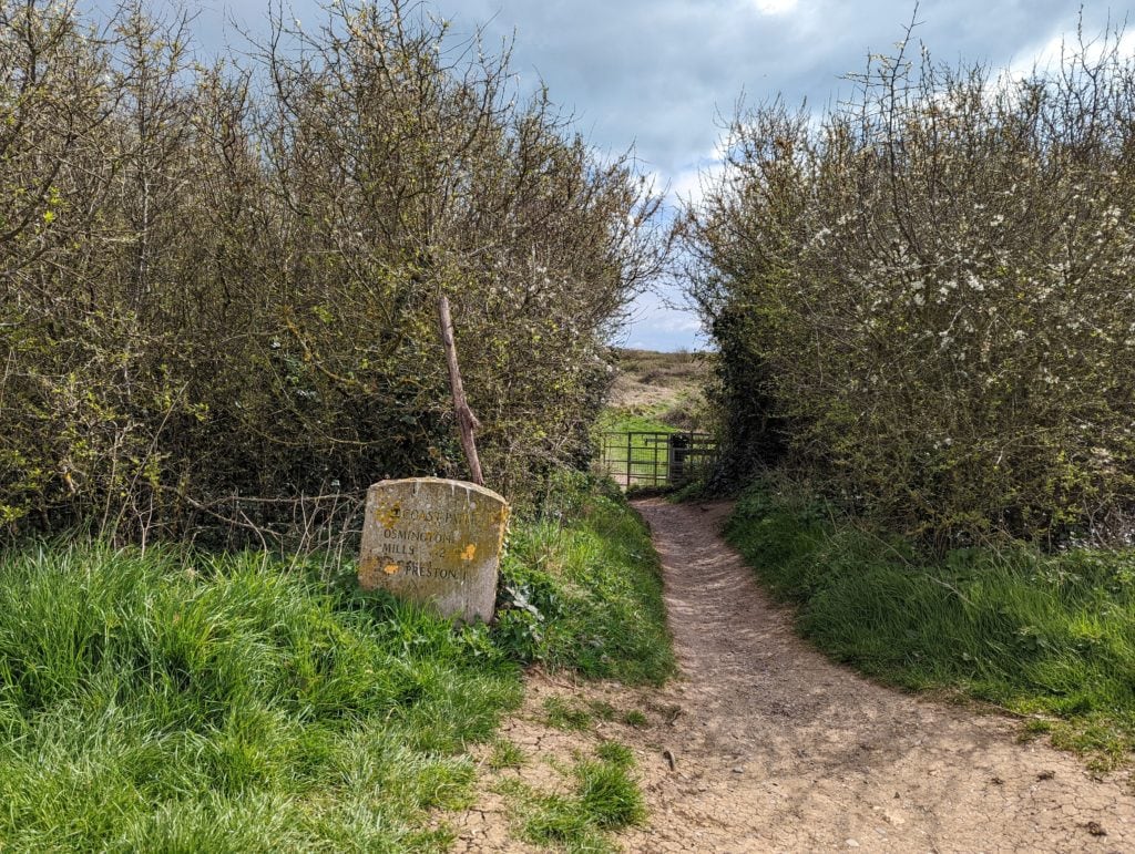 a stone route marker for the coast walk from Weymouth to Osmington Mills next to a dirt path leading twoards a metal gate between two hedgerows