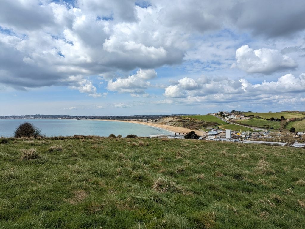 looking downhill towards a cove with a sandy beach and a collection of buildings, there is a hill in the background with houses on it