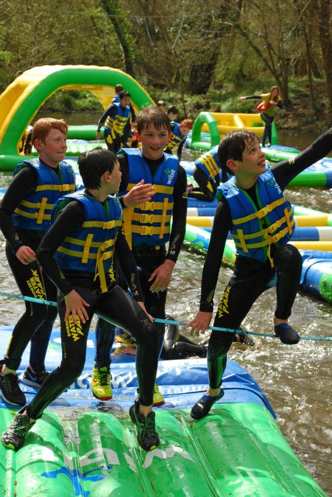 4 boys wearing black wetsuits and blue life jackets with yellow straps are all standing on a green inflatable pad on top of grey water in a lake - dorset adventure park is one of the best places in dorset to visit with older kids. 