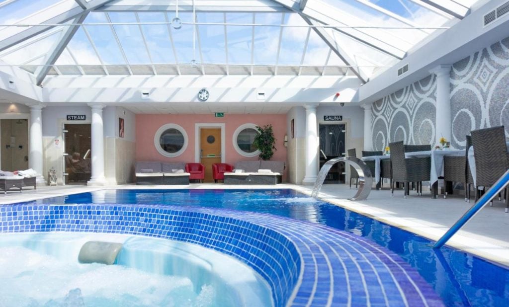 the edge of a hot tub surrounded by a blue tiled circle with a swimming pool behind. all inside a large conservatory style building with a glass roof. one of the best hotels with spas in dorset.
