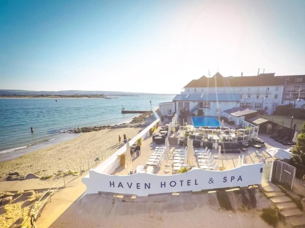 a sandy beach with a hotel on the edge of the sand and next to the sea. there is an outdoor swimming pool next to a seating area with a large sign saying haven hotel and spa. one of the best hotels with spas in dorset. 