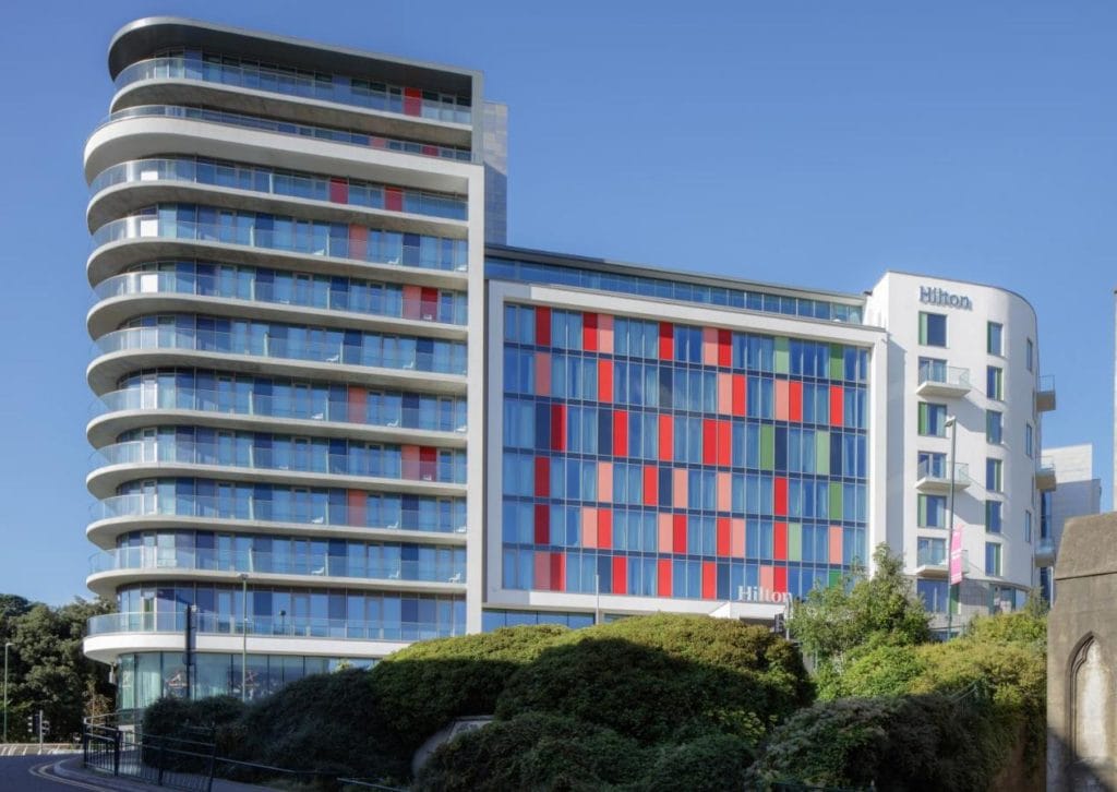 a tall hotel building with a Hilton sign at the top. It isabout 15 storeys high and uilt mainly from glass. The sky behind is blue. 