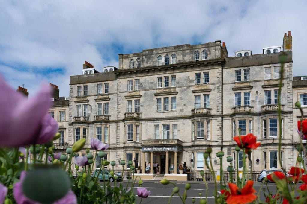 looking through purple and red flowers towards one of the seafront hotels in weymouth, a large grey stone building with many windows