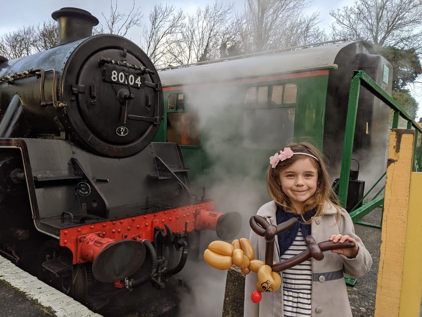 a small girl wearing a cream coloured felt coat and a white and blue striped dress with pink flowers in her hair holding a brown balloon animal. she is standing in front of a black steam engine.