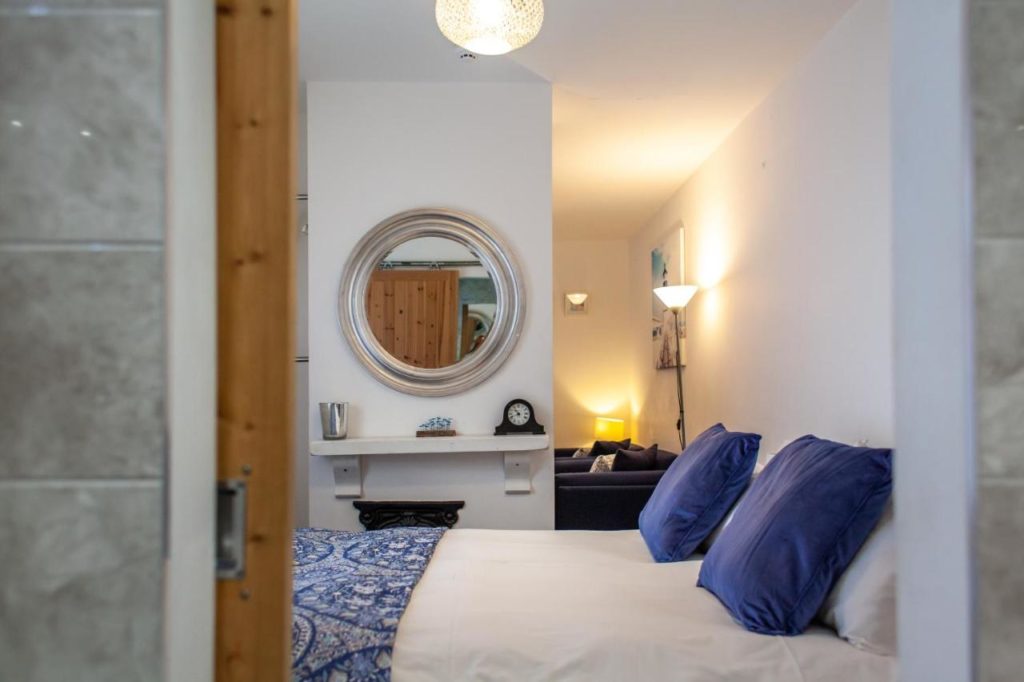 a hotel room with a bed with white bedsheets and blue cushions with a white wall and a round silver framed mirror on the wall
