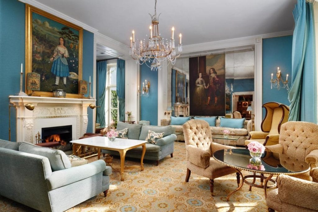 a living room with a patterned biege carpet and light blue walls filled with lots of antiques and furniture and a glass chandelier hanging from the cieling. both walls have large old fashioned paintings on them.