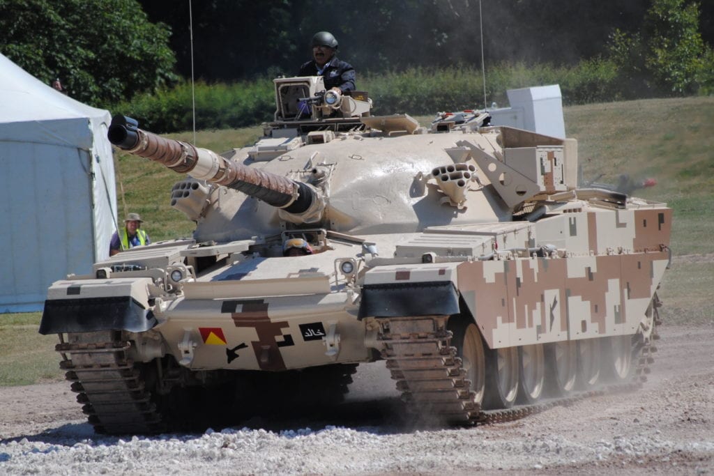 a man in a helmet is sticking his head out of a large grey and brown tank driving on a gravel road at bovington tank museum in dorset