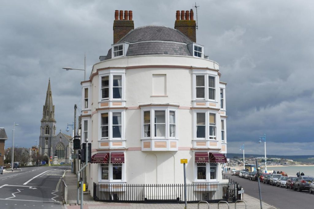 a three storey white painted hotel with bay windows and a slate grey roof on the end of weymouth seafront with a beach on the right side and a church in the background