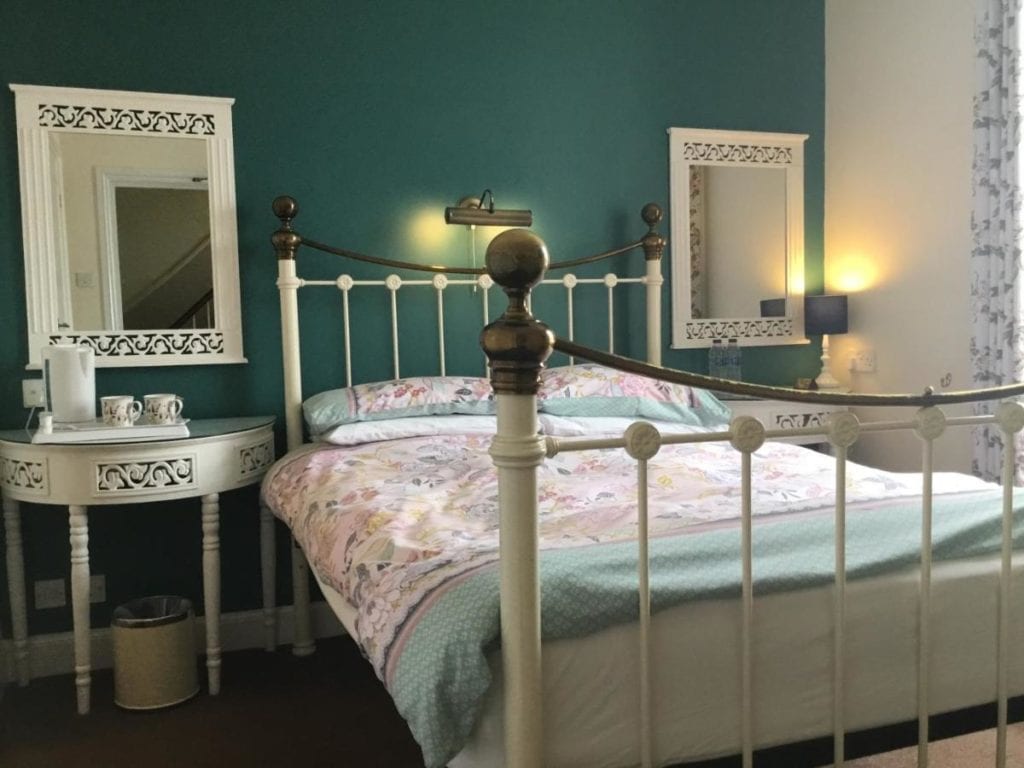 a hotel room with a green wall and a white painted iron bed with pale green and pink sheets next to a small white bedside table