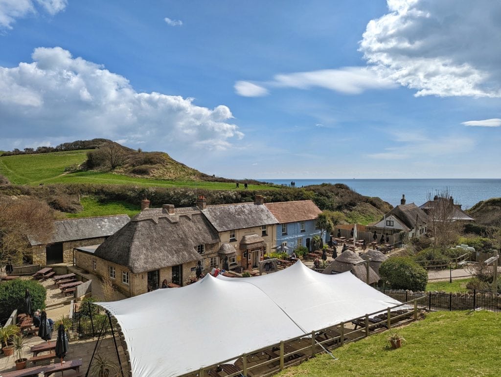 a row of cottages painted yellow and blue with thatched roof in front of a small cliff with the sea behind and a large white marquee in front all forming the Smugglers Inn pub in Osmington