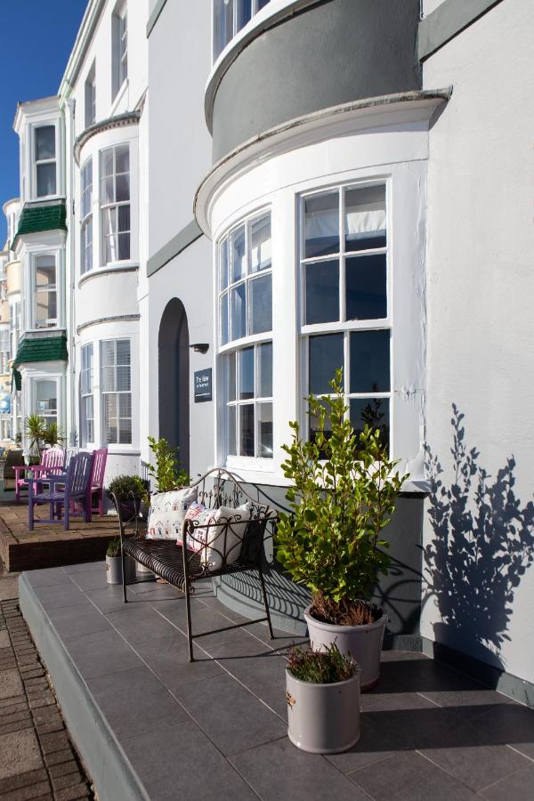 two narrow three storey hotels painted grey with curved bay windows on weymouth seafront with an iron bench and some pot plants outside