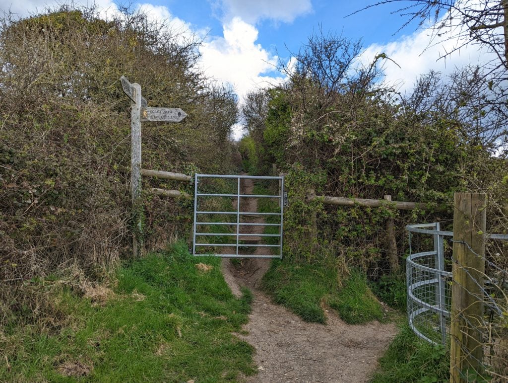 a metal gate between hedgerows with a wooden signpost for the south west coast path from weymouth to osmington 