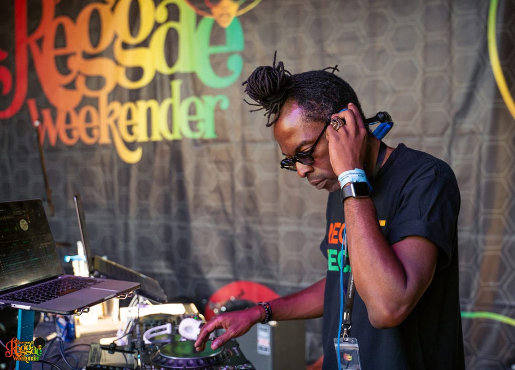 black man with dreadlocked hair in a knot on top of his head wearing a black t shirt and sunglasses and holding headphones to one ear whilst DJ-ing on decks on a stage with a grey curtain behind with the text 'Bournemouth Reggae Weekender in multicoloured letters