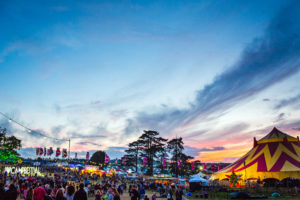 just after sunset with a golden glow at the base of a dark blue sky above a crowd of people and a circus tent at Camp Bestival, one of the biggest music festivals in Dorset