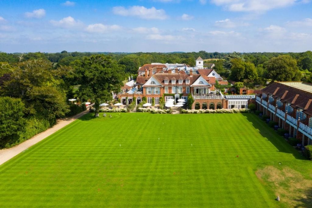 drone shot of a large green field with a big red brick mansion at the far side and green woodland behind that