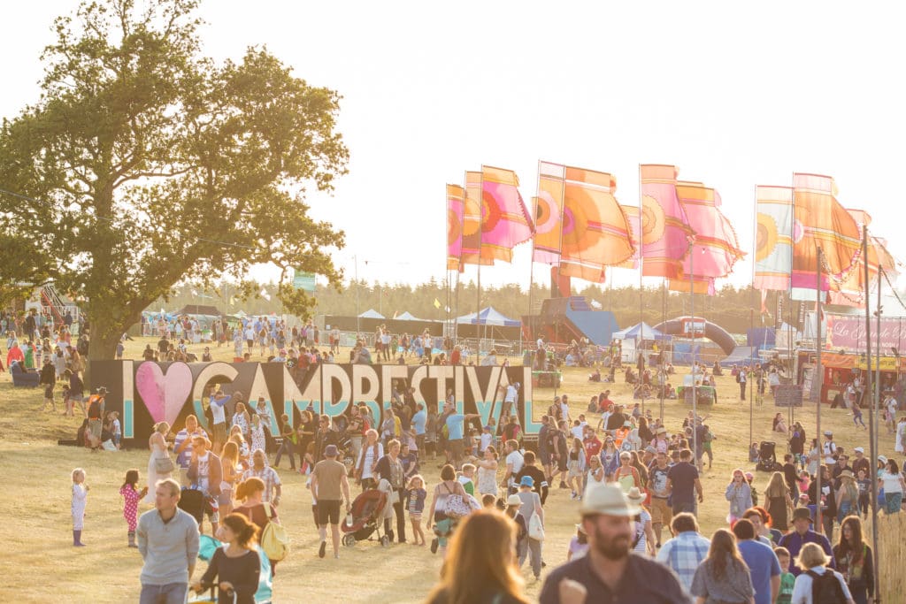 a large sign in a field reading I heart Camp Bestival surrounded by people and several waving pink flags at sunset with everything turned golden by the setting sun. this is one of the biggest music festivals in Dorset.  