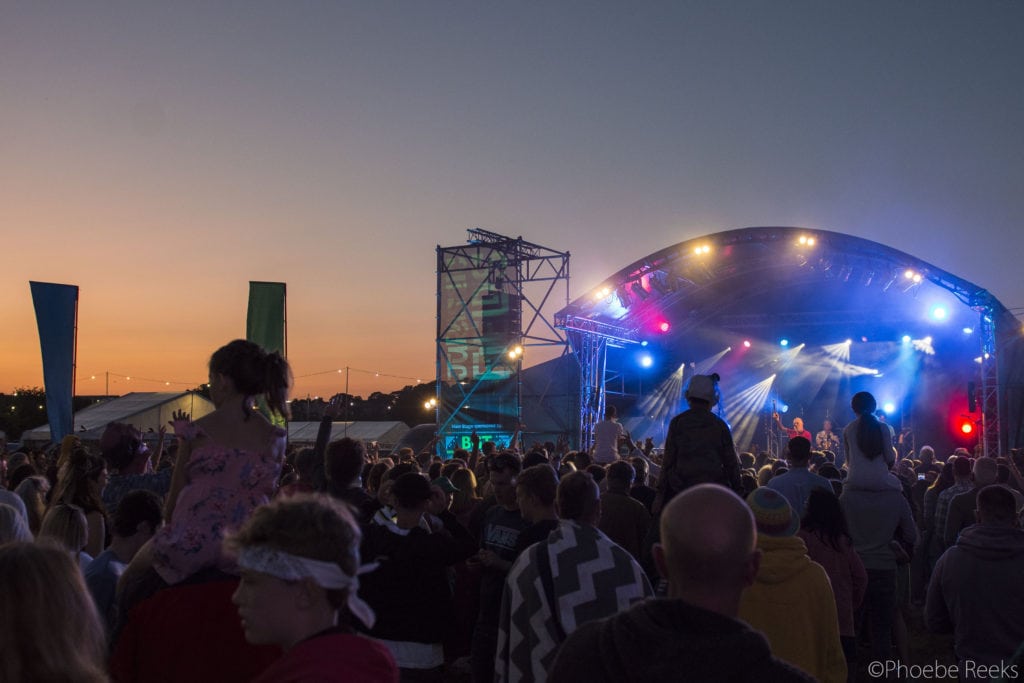 a lit up stage with a crowd in front silhoutted against a dark orange sky just after sunset