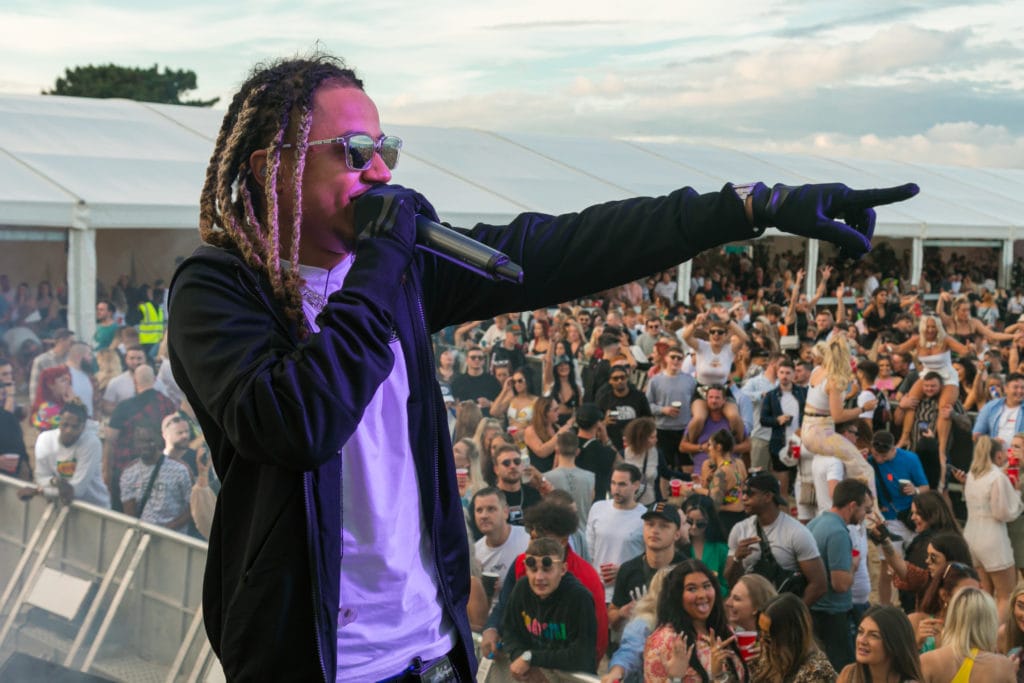 a man with dreadlocks wearing sunglasses is standing on a stage in front of a large crowd holding a microhphone and pointing at something off camera