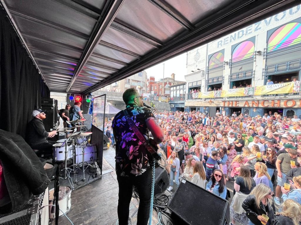 a man on stage in a flowery shirt holding a guitar and singing into a microphone with a large crowd in front of him and a white-painted building behind them