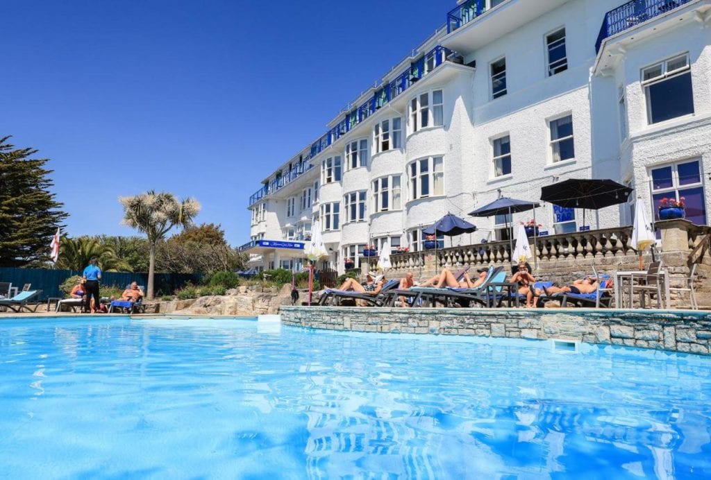 outside of a large white hotel on a very sunny day with a blue outdoor swimming pool in front and a blue sky overhead - dog friendly hotels dorset
