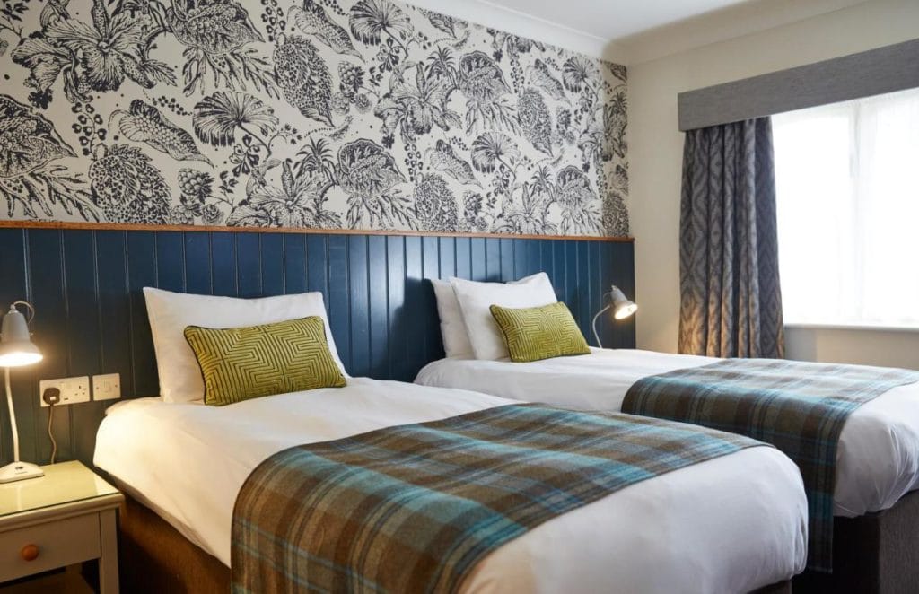 two hotel beds side by side with white sheets and a blue tartan throw over them against a wall with blue wood pannelling and a black and white flower patterned wallpaper above the panels