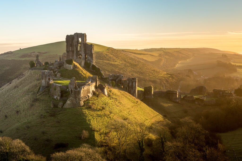 a ruined stone castle on a hill at sunrise with golden light falling on the surrounding grassy hills - corfe castle purbeck dorset