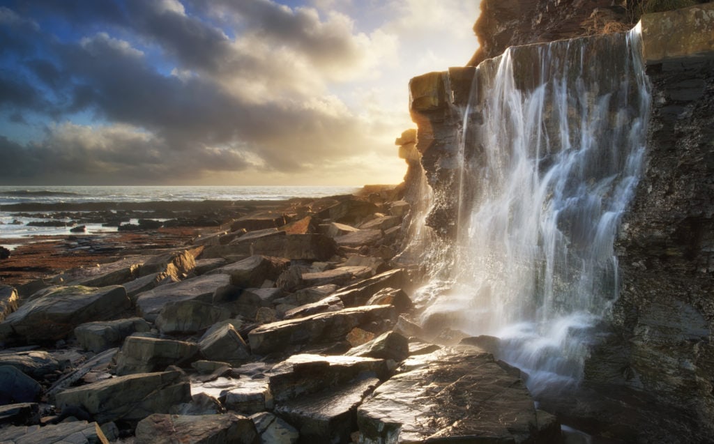 waterfall flowing down a cliff face onto rocks on beach at sunset in kimmeridge bay dorset