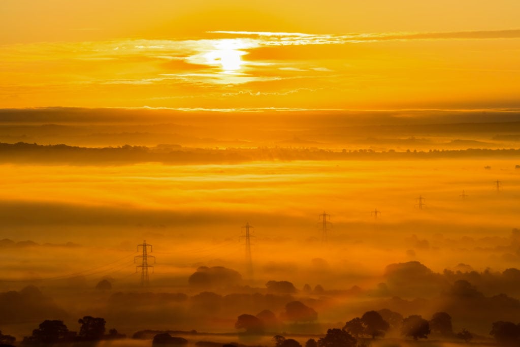a bright orange sunrise sky over a foggy landscape with a few trees and some pylons emerging from the fog and dark orange clouds in the sky beneath the sun 