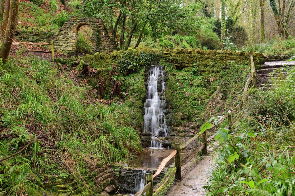 Long exposure of a small narrow waterfall flowing down a stone wall covered in moss next to a path in Ninesprings park in Yeovil in Somerset