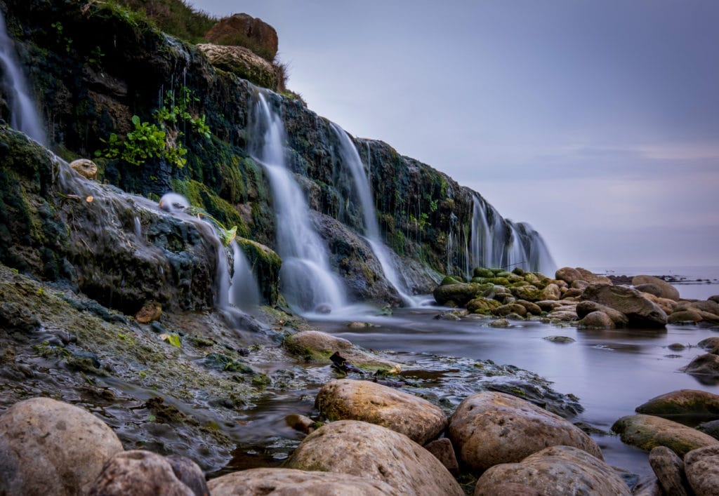 long exposure of a very small waterfall flowing down a rock face into a pool of water on a stoney beach just around sunset with a purplish sky at osmington mills in dorset