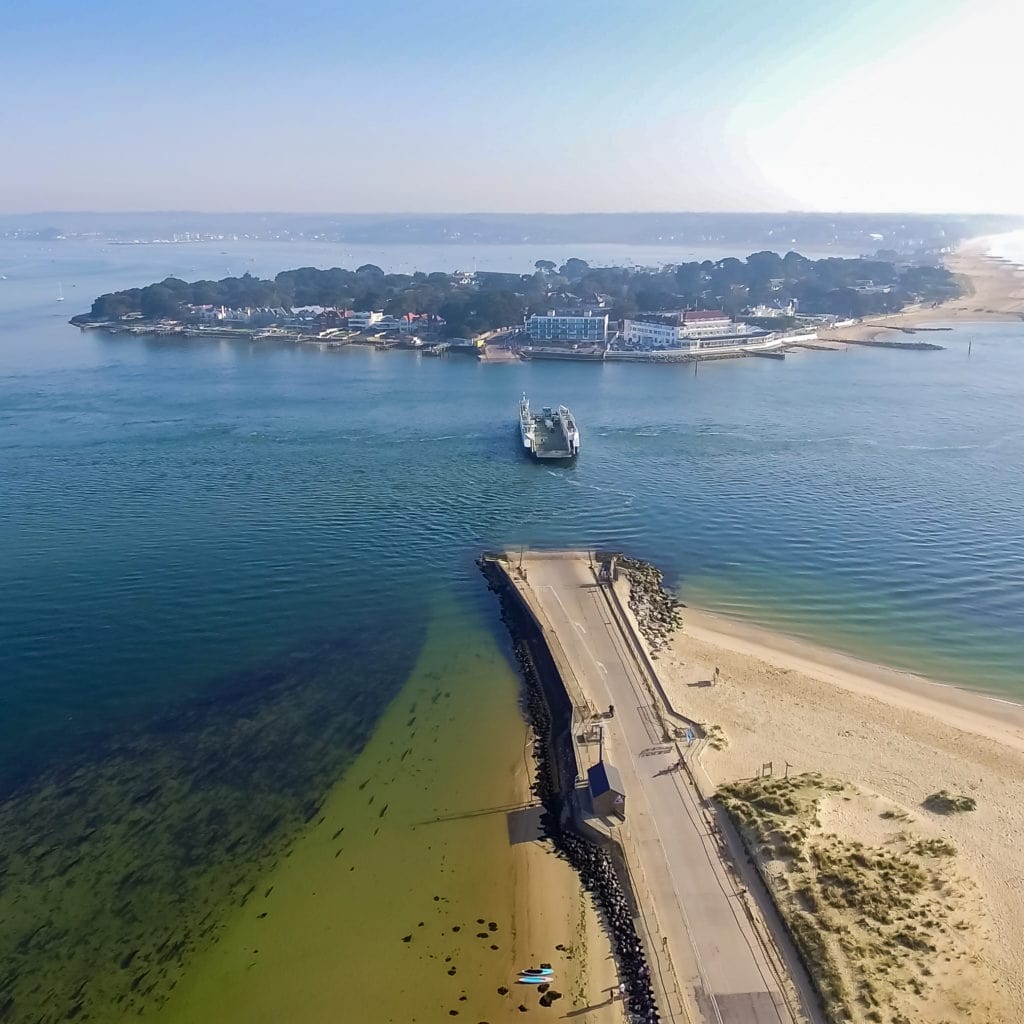 drone shot of a sandybeach with a long road stretching across it towards the sea. there is a ferry in the sea crossing a narrow gap from a sandbanks peninsula in the background towards the beach at studland on a sunny day. things to do near bournemouth. 