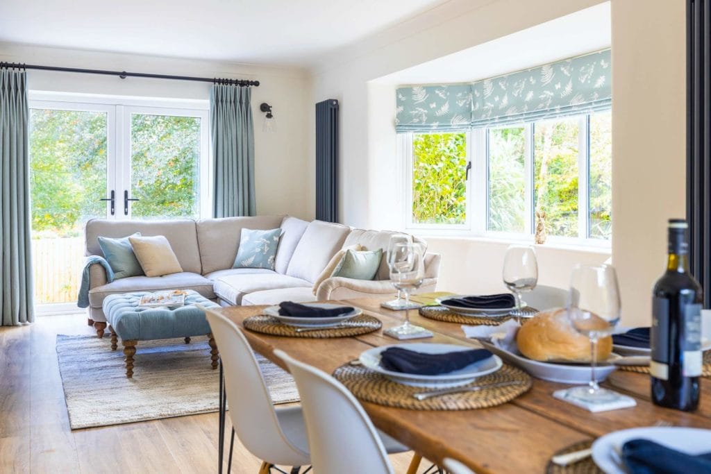 interior of a dining room with white walls a large window and french doors on a sunny day with lots of light. there is a wooden table in the foreground laid with blue plates and cutlery and a light grey sofa behind it. 