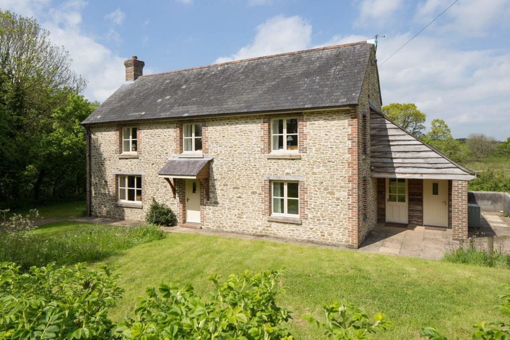 a large stone holiday cottage in dorset with a grey slate roof and light beige stone with three windows upstairs and two downstairs and a white front door on a sunny day with a grassy lawn in front. 