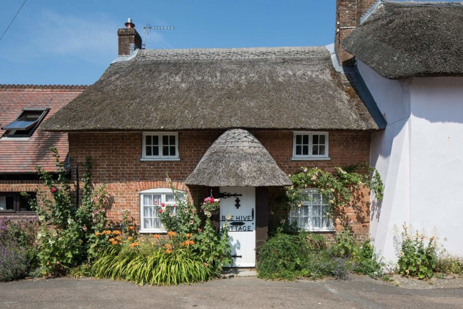 red brick cottage covered in flowering climbing plants with white framed windows and a white front door and thatched roof on a sunny day with clear blue sky.