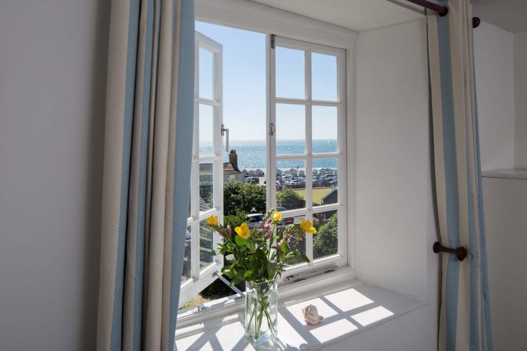 open windw in white bedroom walls with grey curtains looking out onto a green garden with a small town behind and the sea behind that. there is a glass vase with daffodils on the windowledge.  