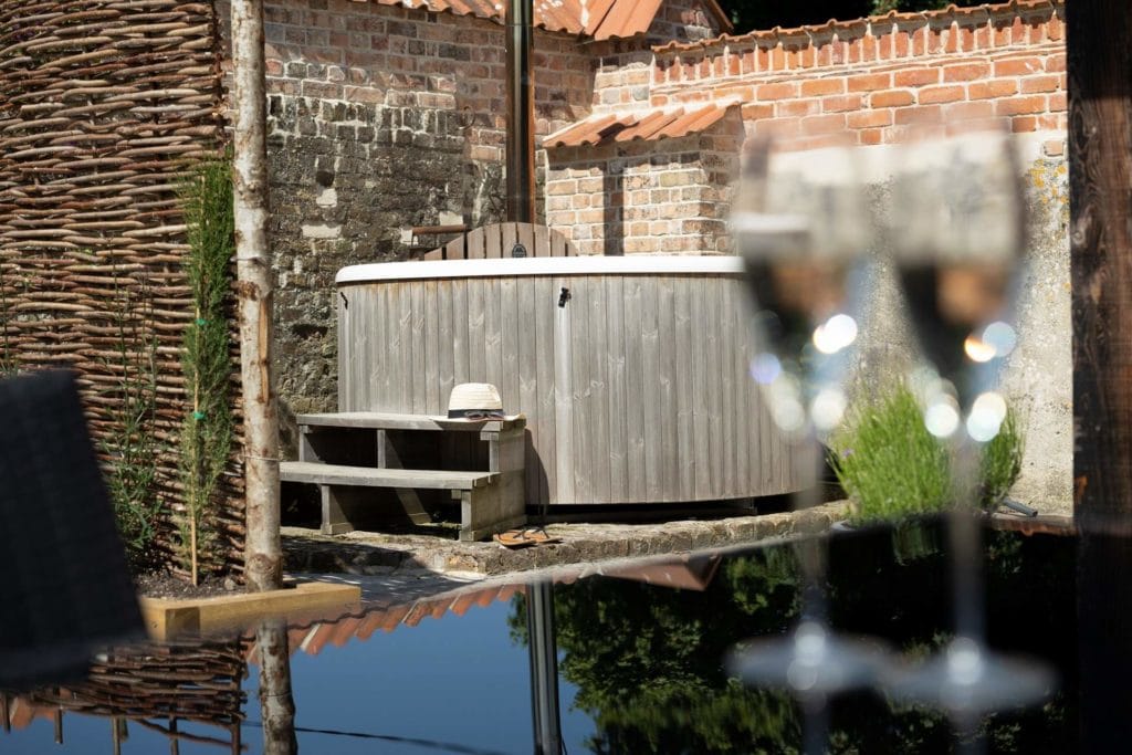 two champagne glasses out of focus on a table in the foreground with a wooden outdoor hot tub against a stone wall behind outside on a sunny day