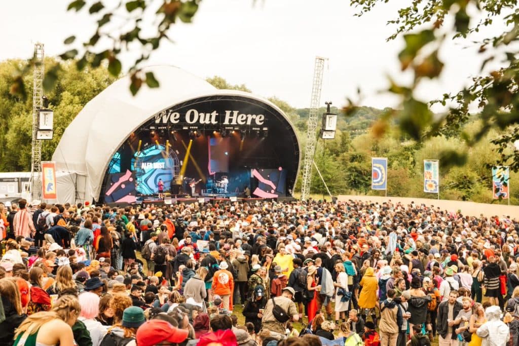 oval shaprd stage with a white roof and a huge crowd of people in front surrounded by trees on a sunny summer day