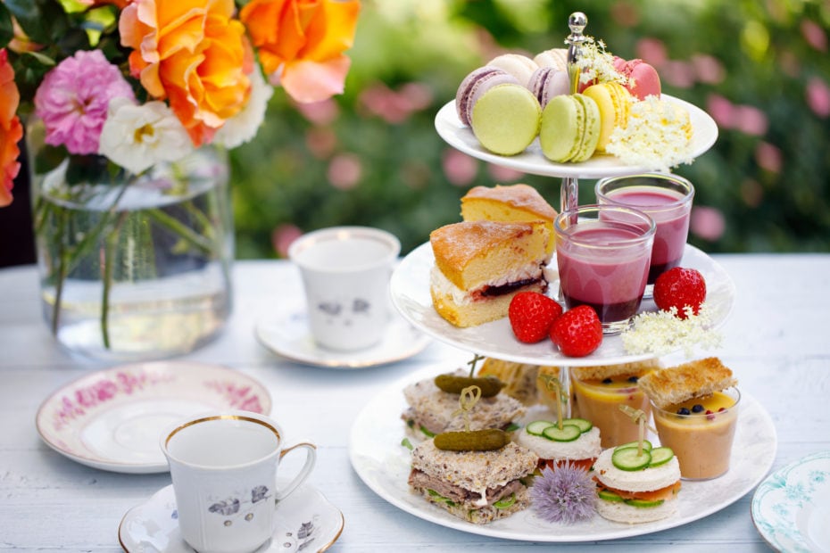table outside in a garden with a white tablecloth with a vase of colourful flowers on it and a three tiered white china cake stand with sandwiches scones and cakes on it and two china teacups alongside. Best afternoon tea in Bournemouth.