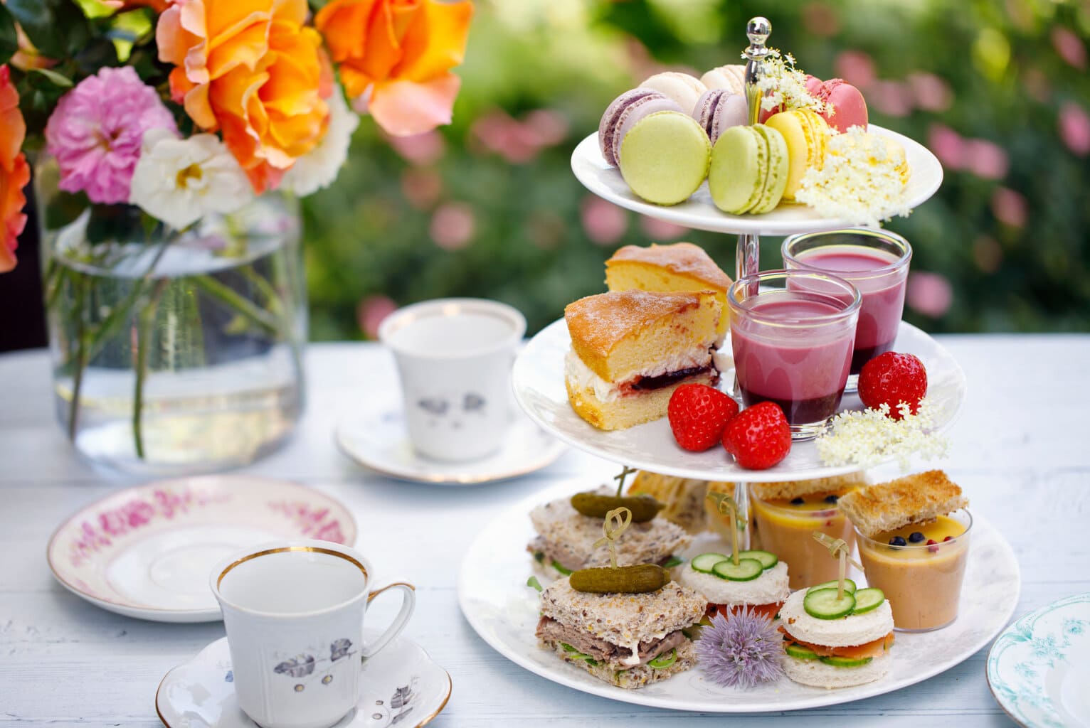table outside in a garden with a white tablecloth with a vase of colourful flowers on it and a three tiered white china cake stand with sandwiches scones and cakes on it and two china teacups alongside. Best afternoon tea in Bournemouth.