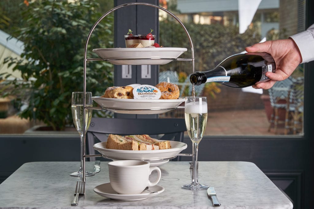 three tiered metal afternoon tea stand with cakes scones and finger sandwiches on a white marble tabletop with two champagne flutes and a hand pouring a bottle of champagne at the The Connaught Hotel in Bournemouth