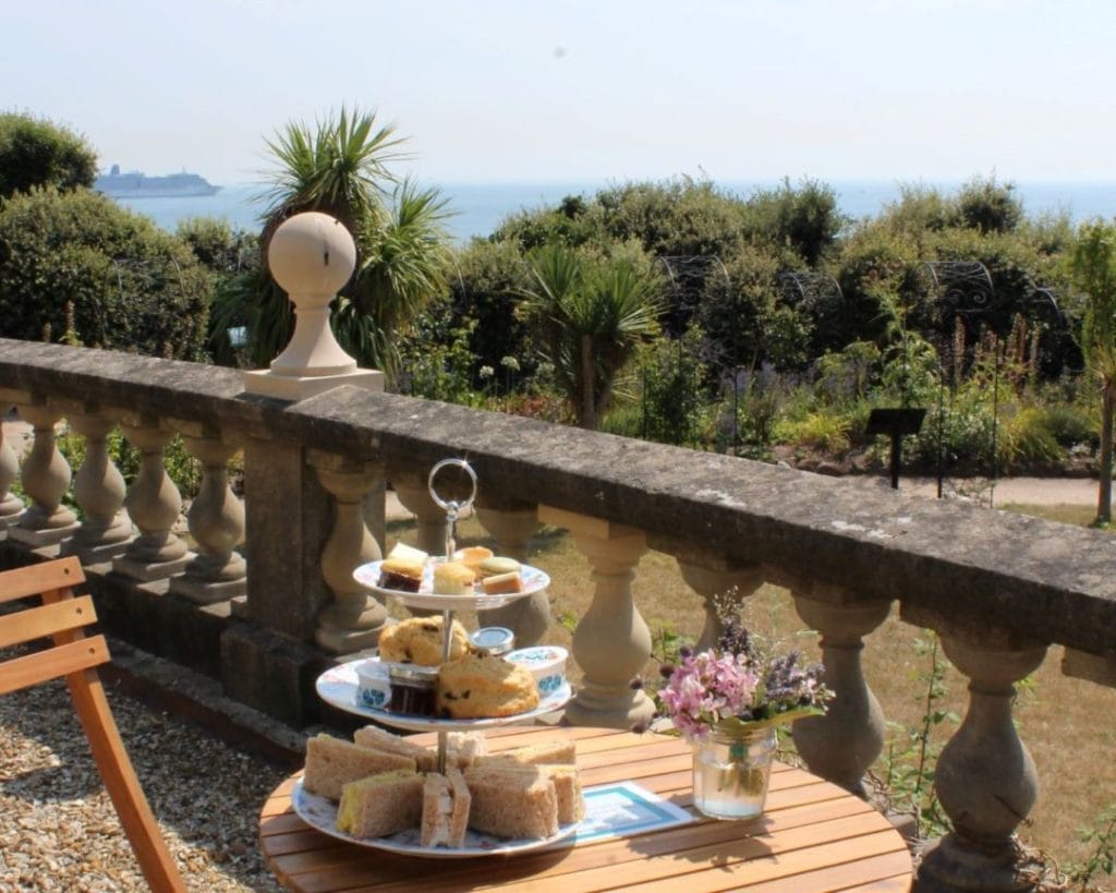 outdoor terrace with a low stone wall above a grassy lawn and green shrubs. there is a small wooden cirtcular table on the terrace with a three tiered white china cake stand on it laid with cakes and sandwiches for afternoon tea and a small vase with pink flowers next to it. 