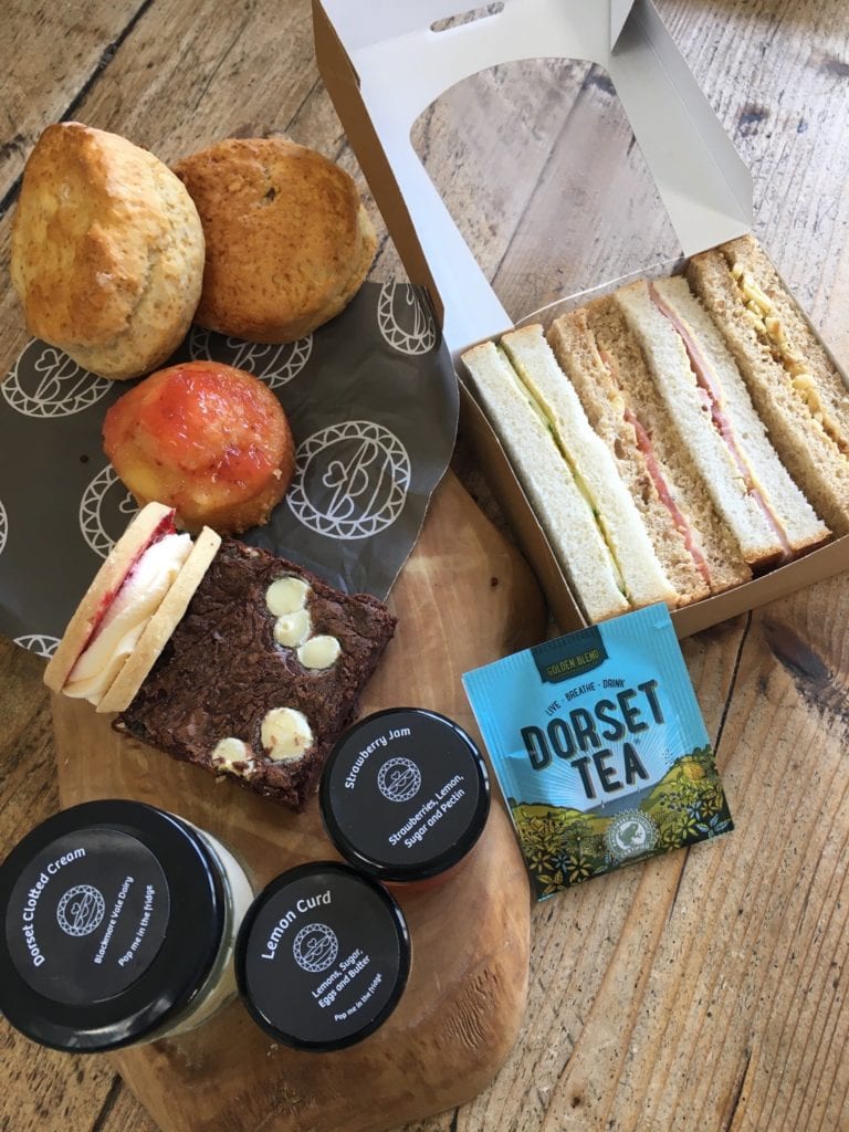 flat lay of a wooden table with a selection of cakes and scones next to a paper box of sandwiches, a dorset tea sachet and three small jars of jams and cream. best afternoon tea in bournemouth. 