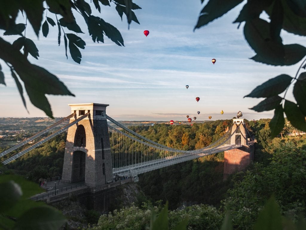 long stone suspension bridge with brown stone towers on either side across a large gorge in bristol on a sunny day with clear blue sky and several colourful hot air balloons floating above