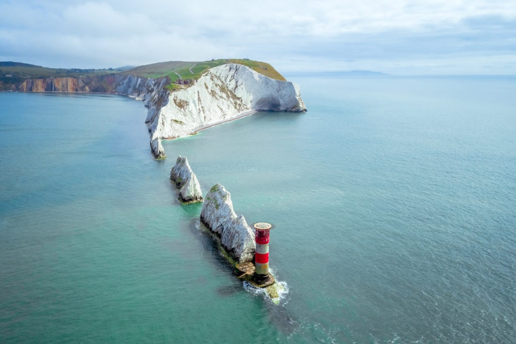 aerial shot of the needles on the isle of wight, a line of triangular shaped white chalk rock stacks leading into the sea from the chalk white cliffs of an island with a small red and white lighthouse on the final rock stack