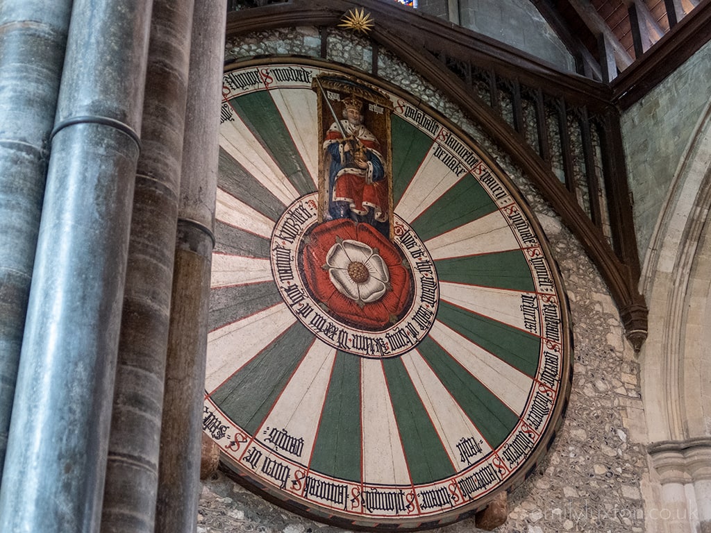 large circular wooden table top mounted on a stone wall in Winchester Cathedral, painted in green and white stripes with a red tudor rose at the centre and a small painting of king arthur at the top