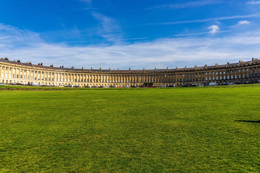 view of the royal crescent in bath, a long crescent shaped row of identical three storey townhouses all built from beige stone with a large empty grass lawn in front and blue sky overhead